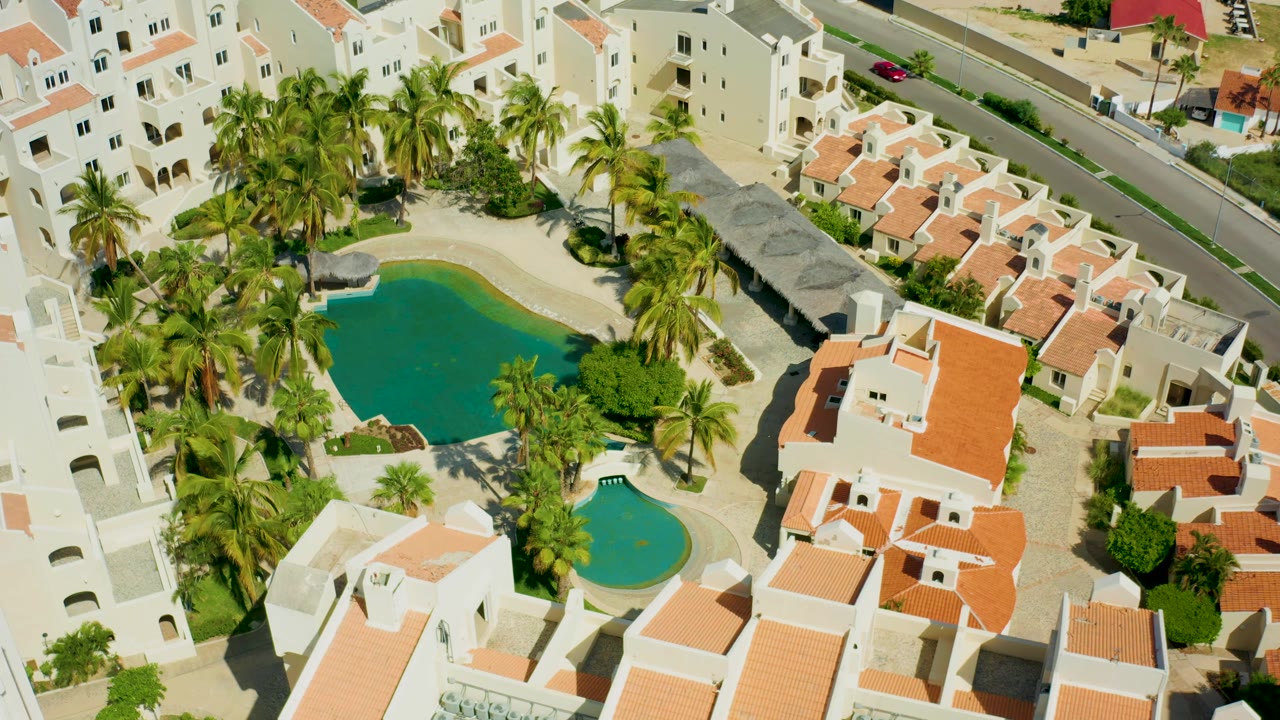 Aerial view over San Jose del Cabo residences, with white villas and orange roofs on the right, palm-lined pools on the left, small cars on the upper road, and no visible people.