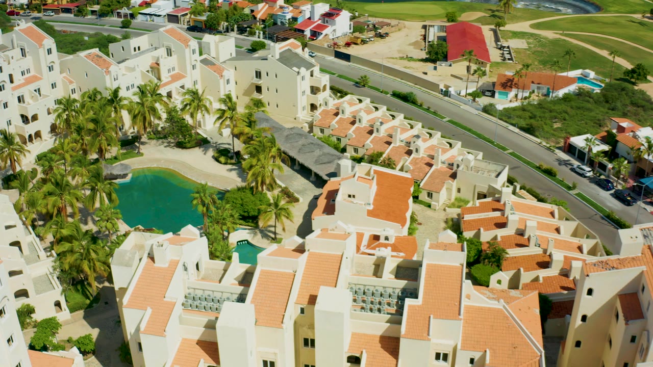 Aerial frame over a San Jose del Cabo residential complex with white buildings, orange tile roofs, a green pool, palm trees, a road at the top of frame, and no visible people.