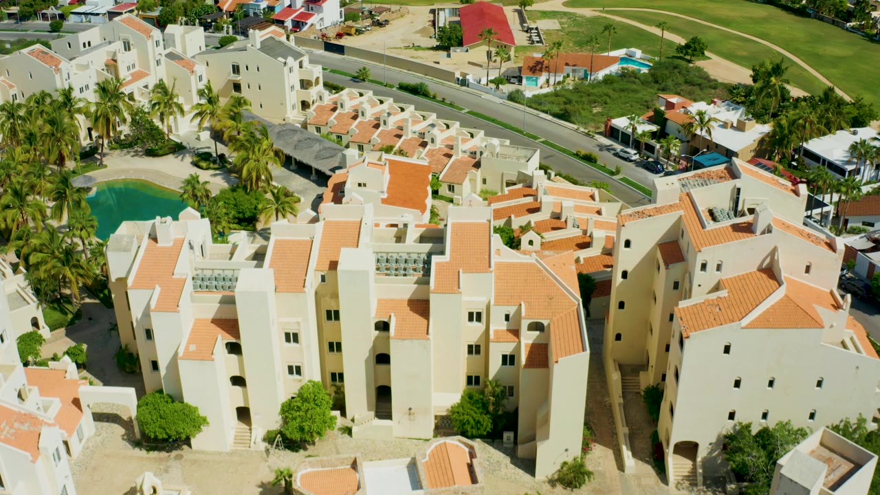 Opening aerial frame over a white residential complex with orange tile roofs, palm trees, a turquoise pool, nearby road, and no visible people in San Jose Del Cabo, Mexico.