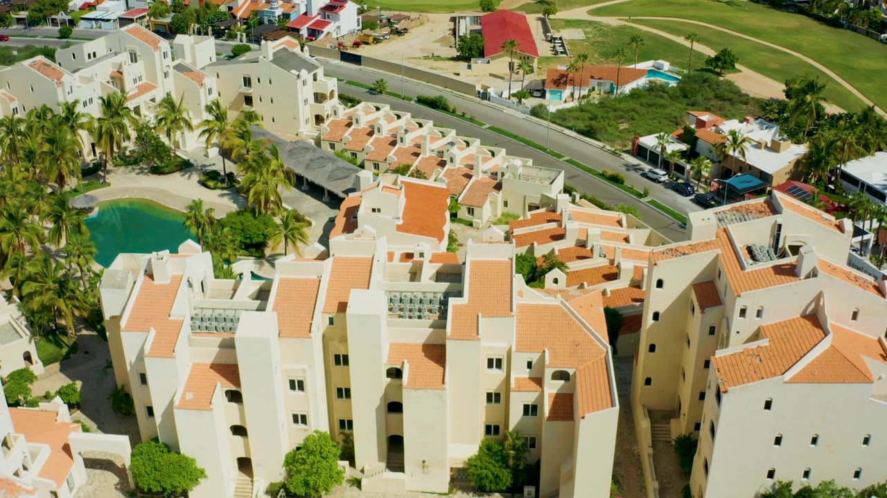 Aerial view of a residential complex with white walls, orange roofs, pool, palms, and road.