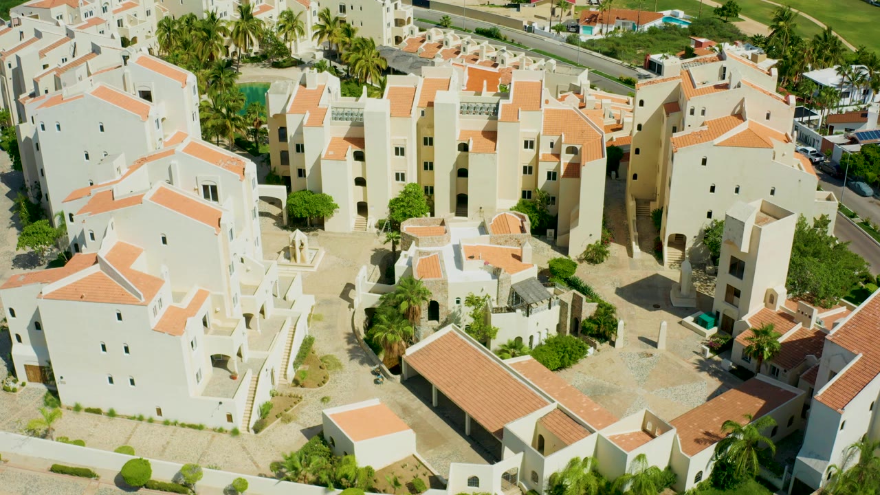 Elevated drone-style view in San Jose Del Cabo showing pale residential buildings, terracotta roofs, courtyard paving, palms, parked cars near the road, and no visible people.