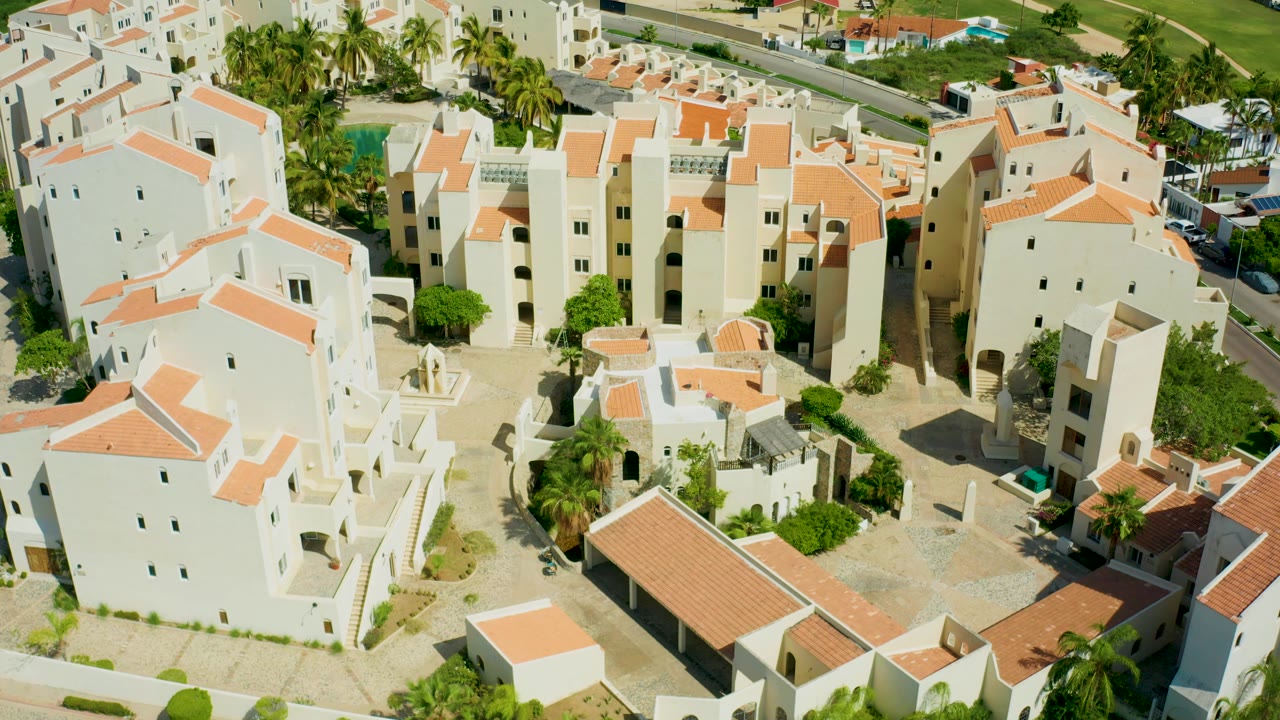 Aerial view of San Jose Del Cabo apartment-style buildings with orange terracotta roof strips, landscaped inner courtyards, palms behind the buildings, and no visible people.