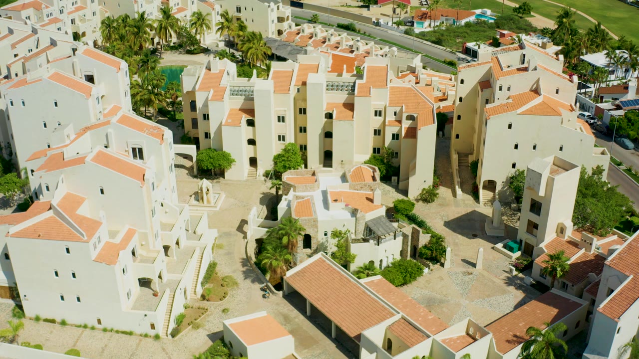 Overhead angle on white buildings, palms, road, and sunlit courtyards.