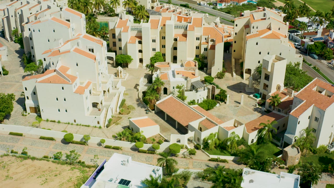 Opening aerial frame over a San Jose del Cabo residential complex with white buildings, red tile roofs, courtyards, palms, sandy ground, and no visible people.