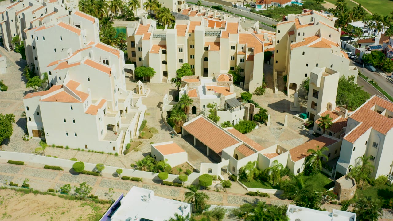 At 0:02, the San Jose del Cabo aerial frames the white residential blocks slightly farther across the roofs, with palms, road strips, sandy ground, and no visible people.