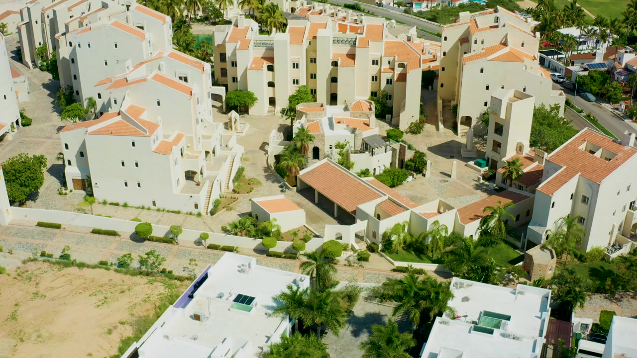 Opening aerial frame over a San Jose Del Cabo residential complex, showing white buildings, orange roofs, dry lots, paved roads, palm trees, and no visible people.
