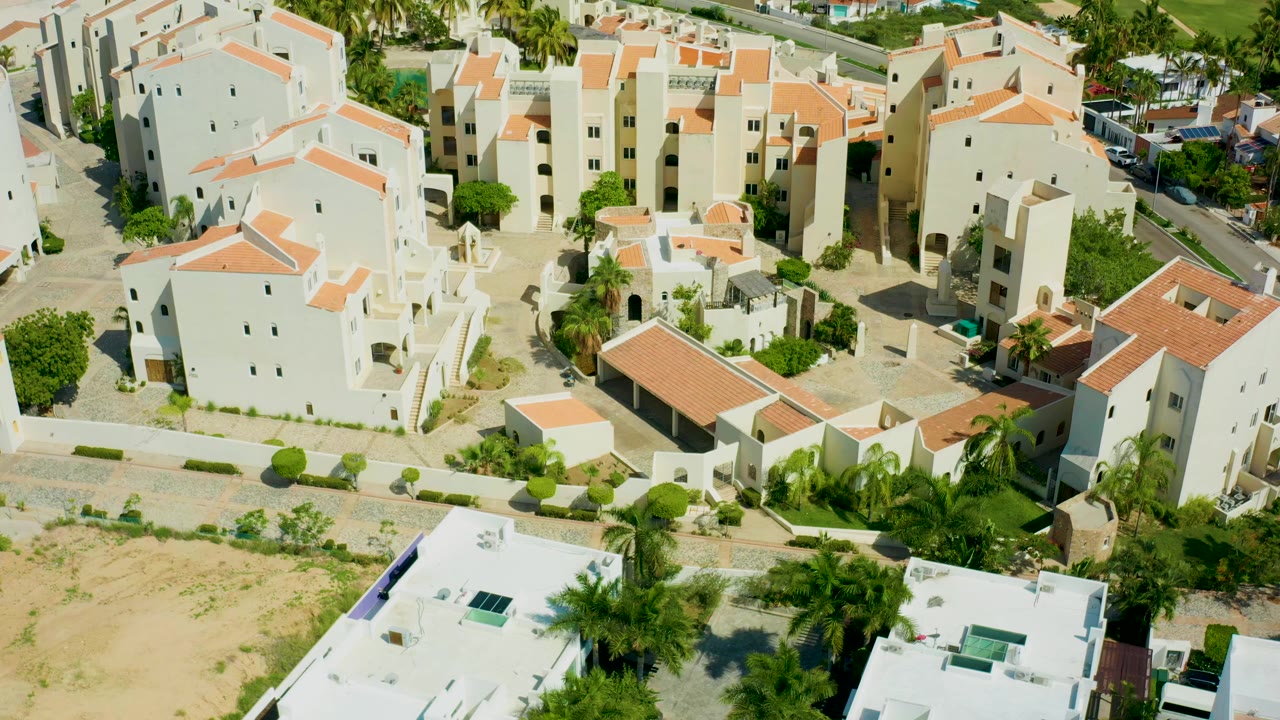 Early aerial moment in San Jose Del Cabo with white low-rise buildings, orange rooflines, courtyards, bordering roads, palm trees, and no visible people.