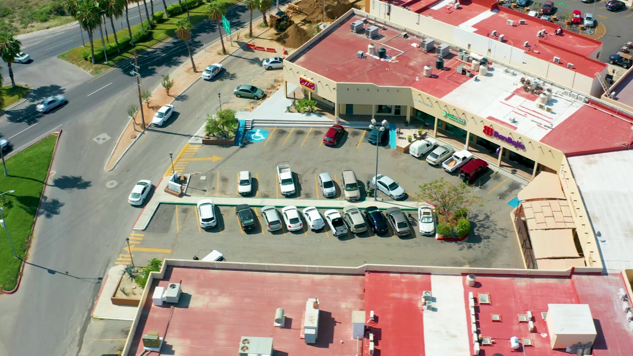 Aerial frame over San Jose Del Cabo with rows of parked cars below red-roofed storefronts, traffic on the road at left, palm trees near the curb, and no visible people.