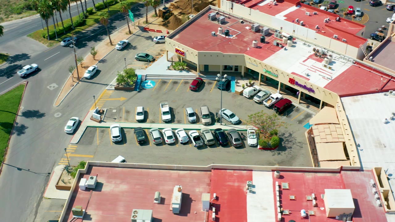 Oblique drone view in San Jose Del Cabo showing the same shopping center parking lot, bright red rooftops, vehicles on the bordering street, and no visible people.