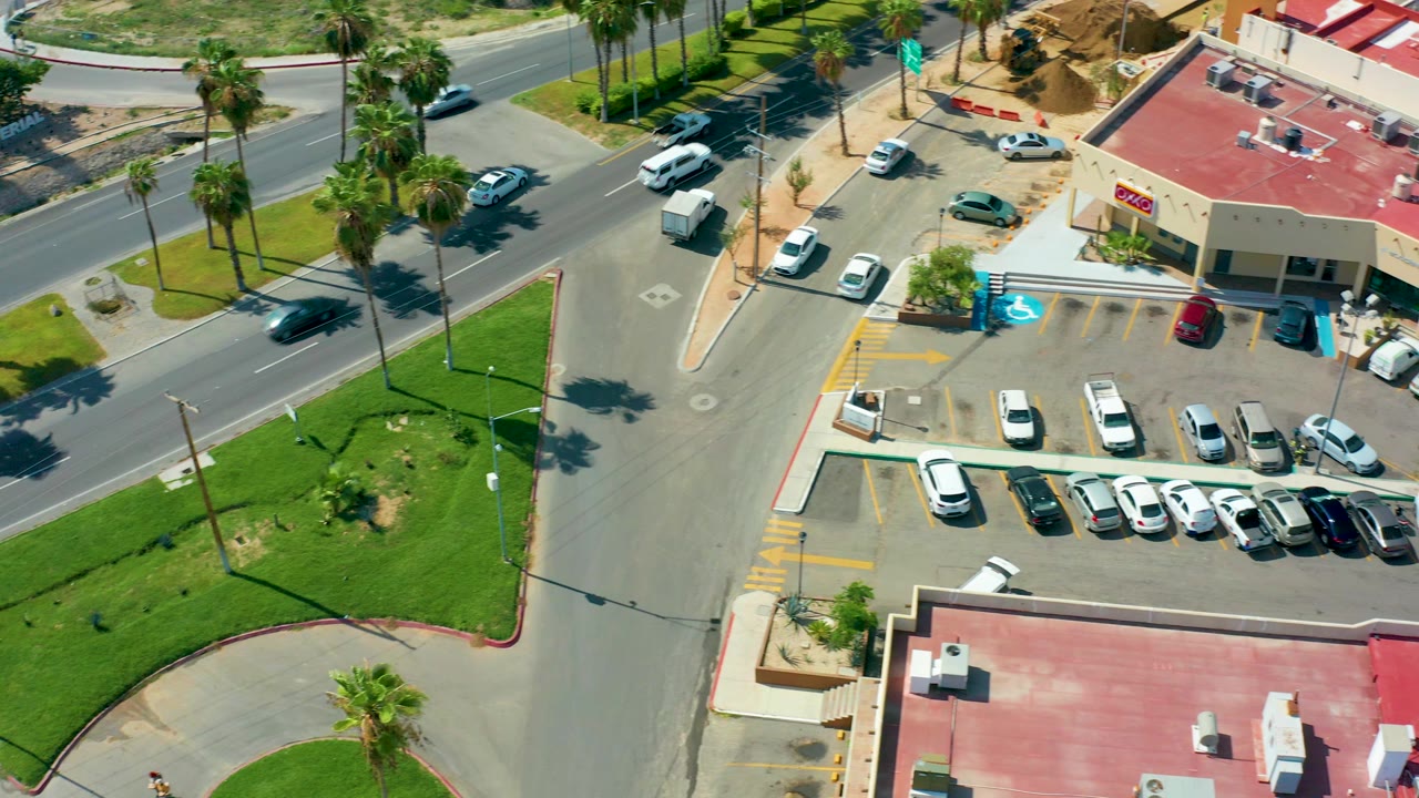High overhead view in San Jose Del Cabo, Mexico, showing a street intersection beside a parking lot, palm trees, grass patches, red-roof buildings, and no visible people at the start of the clip.
