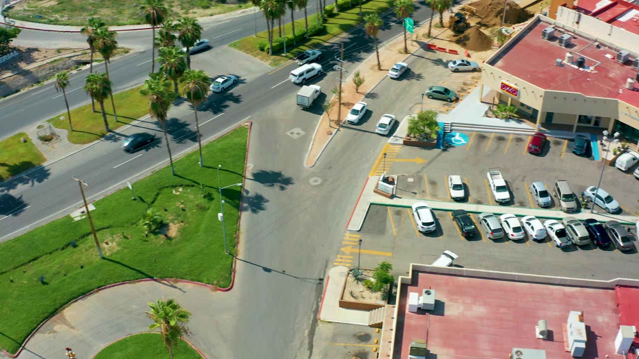 One second into the San Jose Del Cabo clip, the overhead frame keeps the parking lot and road in view as vehicles shift positions along the street, with no visible people.