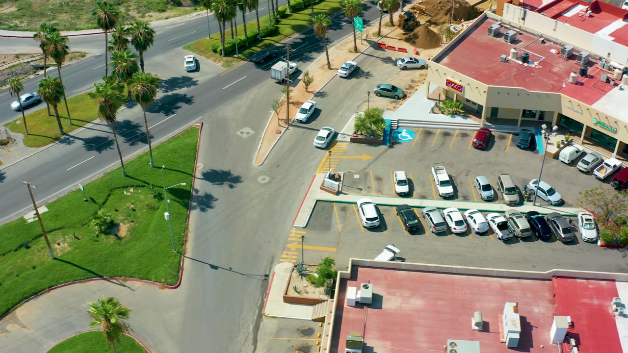Drone-like overhead frame of a street, parking area, palms, and beige buildings with red roofs.