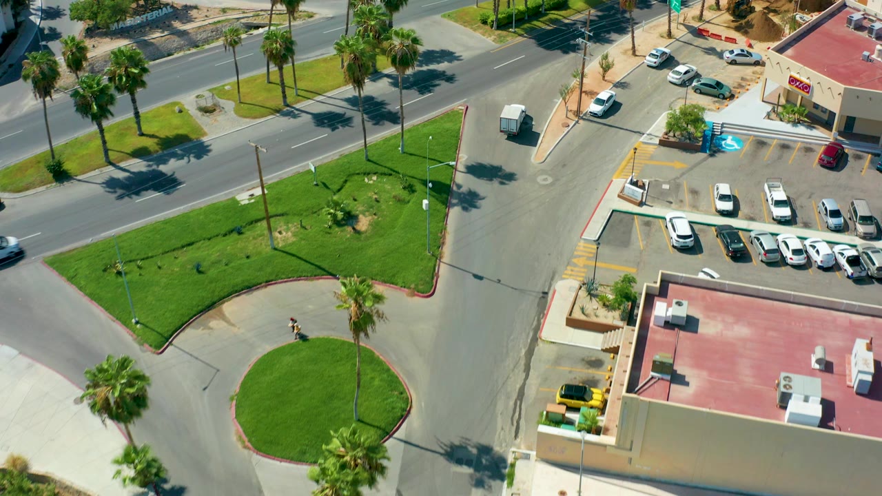 Overhead frame of a San Jose Del Cabo street intersection with green medians, palm trees, parked cars beside a red-roofed building, and no visible people.