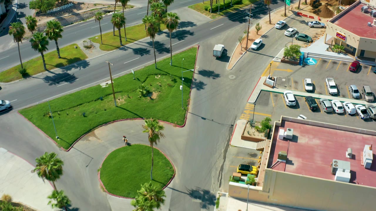 Aerial frame of the San Jose Del Cabo intersection with cars moving along the upper road, a curved green median, palm shadows, and no visible people.