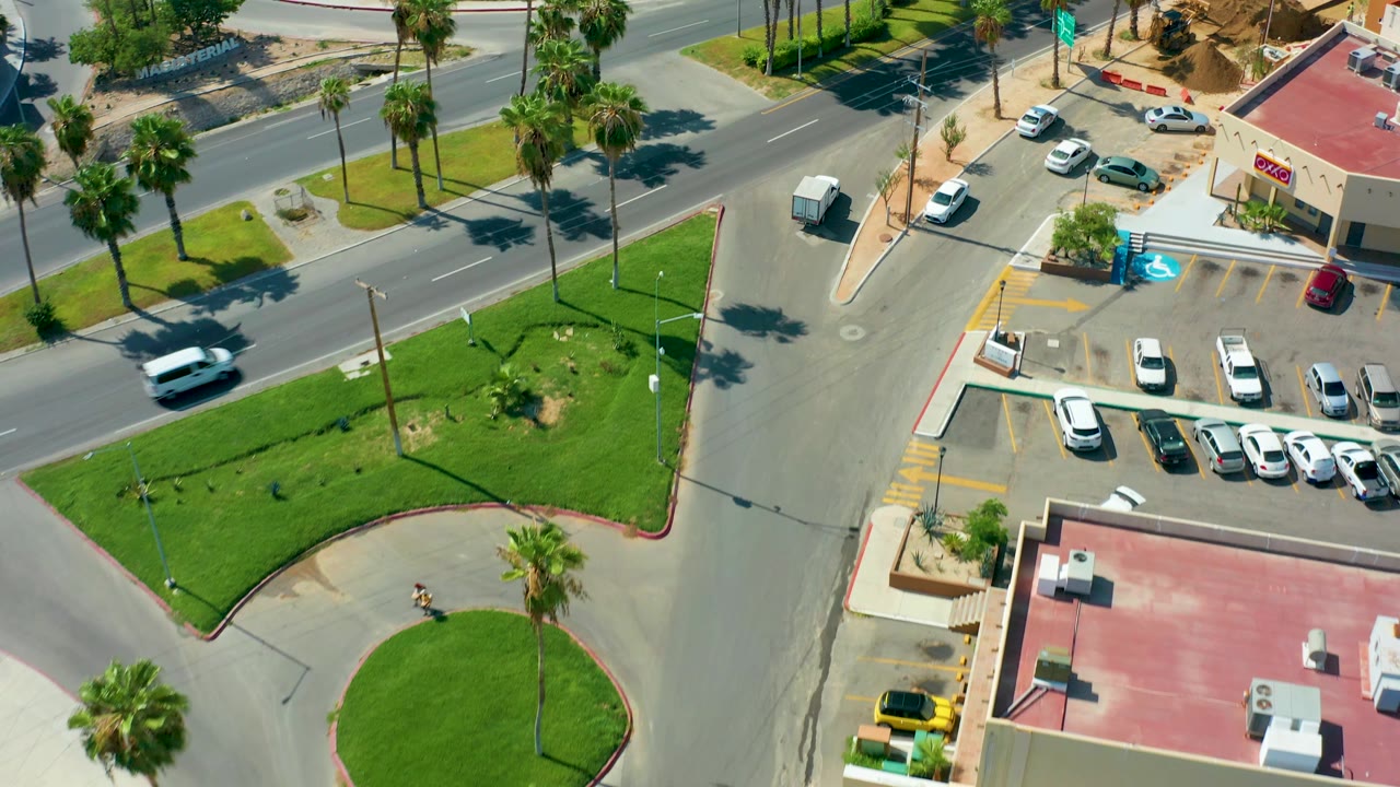 Overhead San Jose Del Cabo street frame with a white vehicle on the road, parked cars in the lot, palm trees along the median, and no visible people.