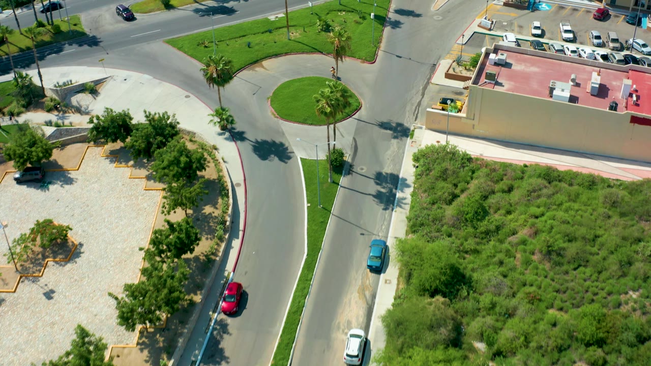 High aerial frame from San Jose Del Cabo, Baja California Sur, showing a rounded traffic island, curved roadway, parking spaces, a red-roofed building, bright lawns, palm trees, and no visible people.
