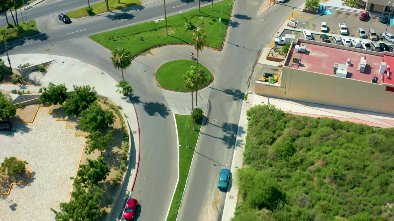 A curved roadway passes between gravel landscaping, brush, and parked cars.