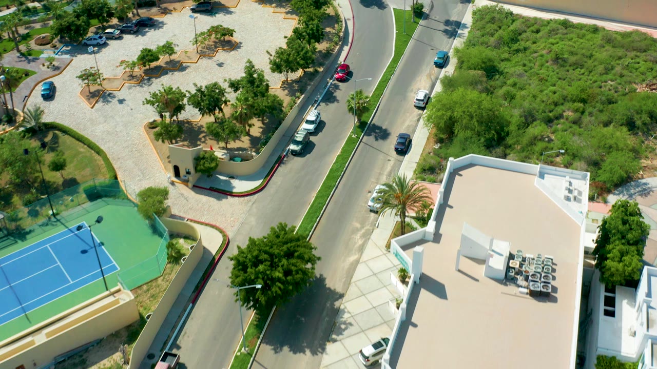 Overhead view in San Jose Del Cabo showing a curved road, blue tennis court, white-roofed building, parked cars, trees, and no visible people.