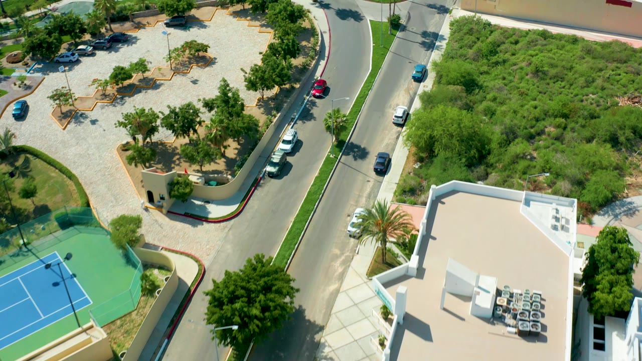 Drone-like overhead frame in San Jose Del Cabo showing the road beside a blue tennis court, a white building roof with rooftop equipment, parked cars, a green undeveloped lot, and no visible people.