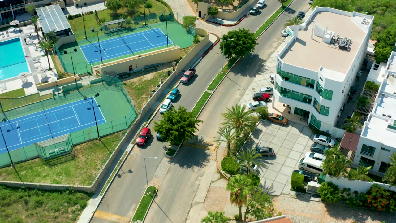 Aerial frame in San Jose del Cabo with the tennis courts centered left, a curved residential road running through the scene, parked vehicles, palm landscaping, and no visible people.