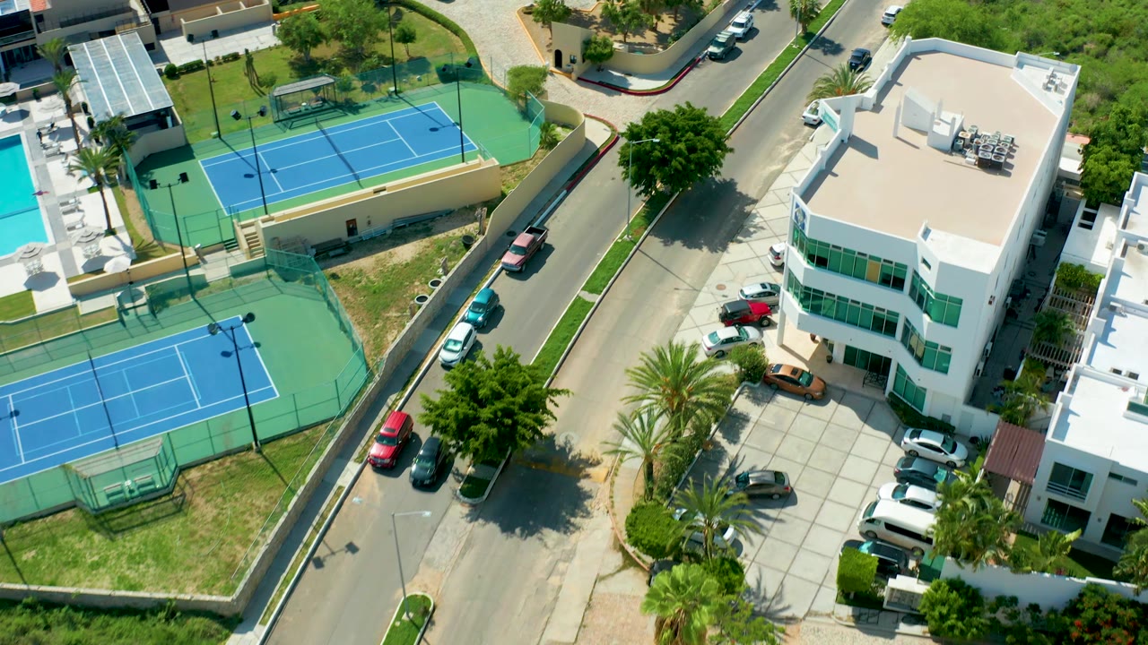 Drone-style overhead frame from San Jose del Cabo showing blue courts on the left, a white flat-roofed building on the right, a residential road, parked cars, trees, and no visible people.