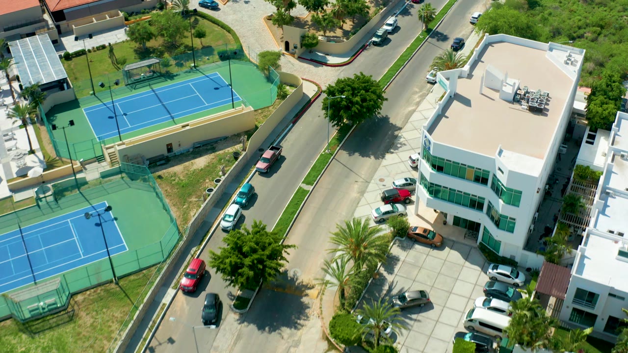 Overhead residential block with tennis courts, parked cars, white building, and palms.