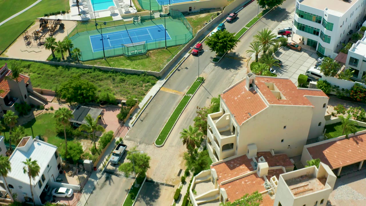 Aerial frame over San Jose Del Cabo shows a road beside blue tennis courts, a pool, residential buildings, parked cars, palms, and no visible people.