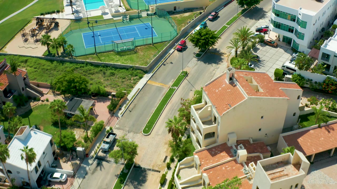 San Jose Del Cabo aerial view shows the road centered between a blue tennis court, pool area, white residential building, parked vehicles, green lots, and no visible people.