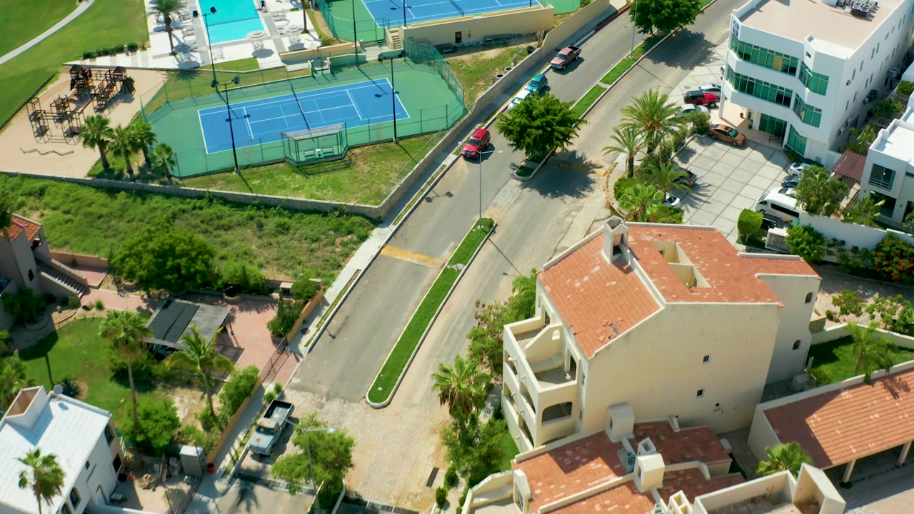 Aerial frame in San Jose Del Cabo shows a red car on the road near tennis courts, a pool, white residential buildings, palm landscaping, and no visible people.
