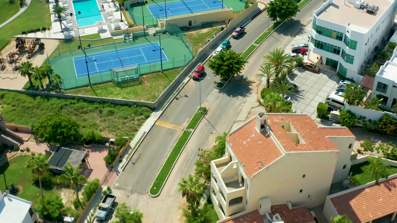 Aerial view of red car on a road near a tennis court, pool, homes, and palms.