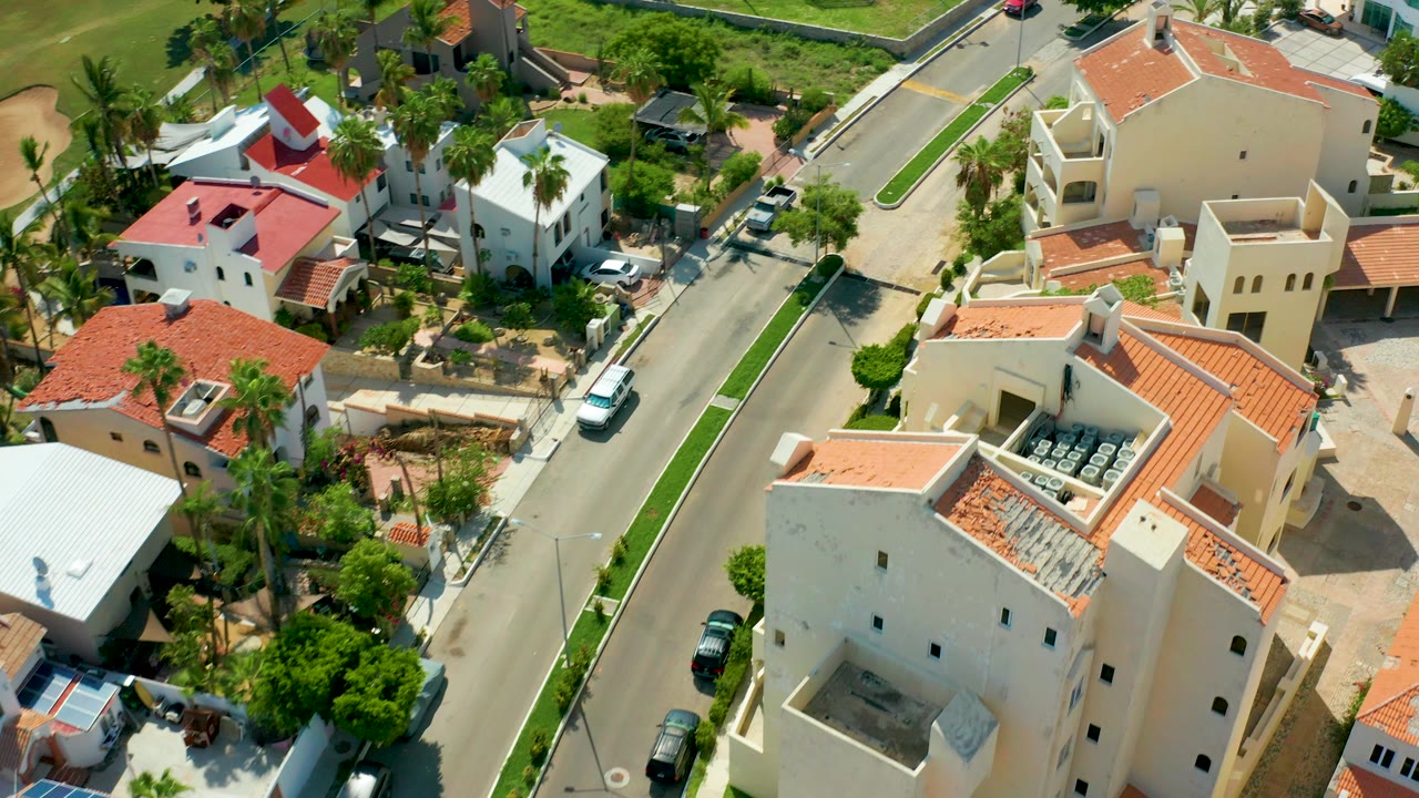 Aerial frame over San Jose Del Cabo homes with red tile roofs, a curved residential street, parked cars, palm trees, and no visible people.