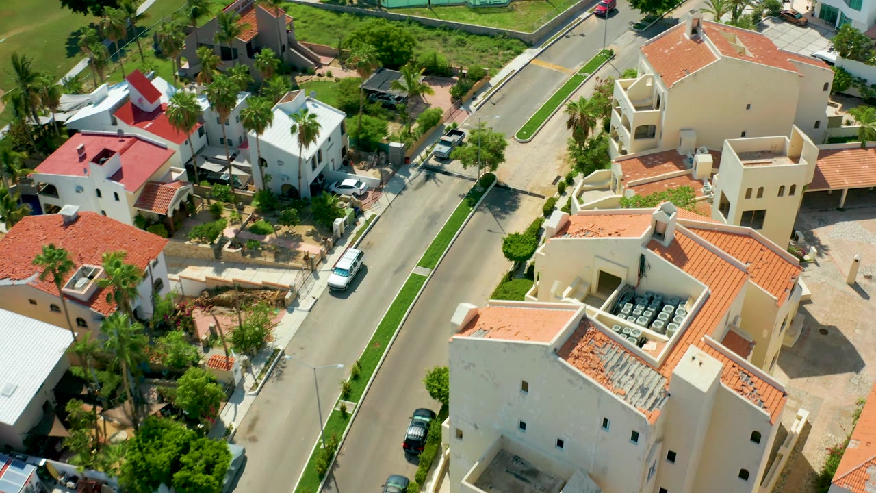 San Jose Del Cabo aerial frame with white homes, red rooflines, a residential road, a white vehicle near the curb, palms, shrubs, and no visible people.