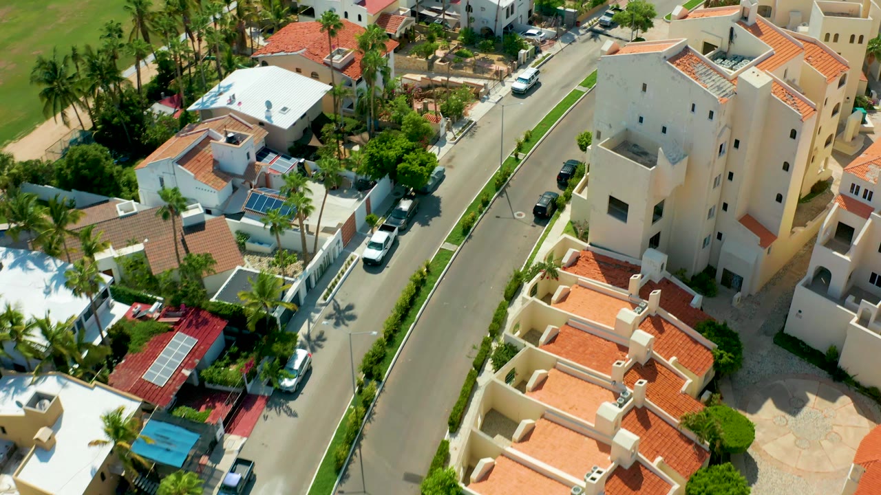 Overhead San Jose Del Cabo frame with the white car progressing along the street, bordered by white and cream houses, palms, red roofs, parked cars, and no visible people.