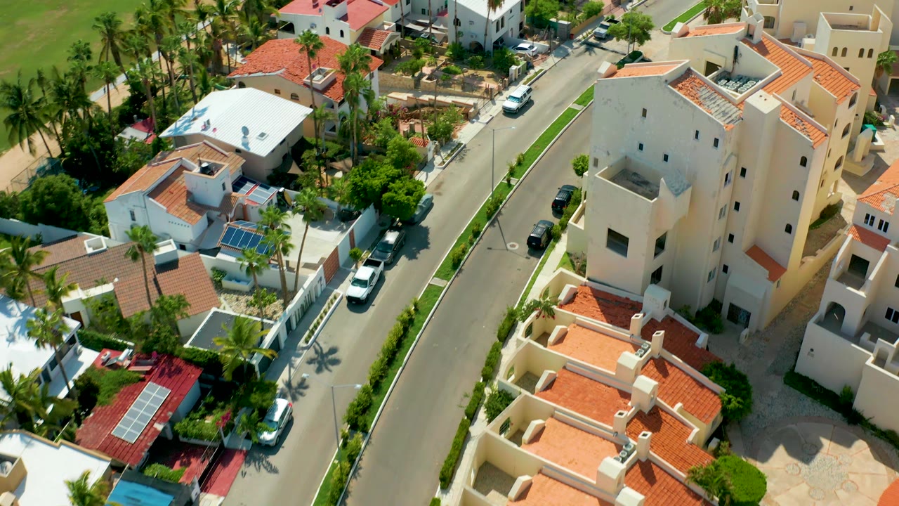 High drone view in San Jose Del Cabo with the white car near the upper road, a large cream residential building, red tile rooftops, palms, parked cars, and no visible people.
