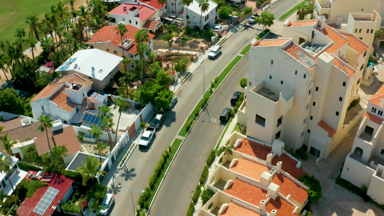 Overhead view of a white car between houses, palms, and red roofed buildings.