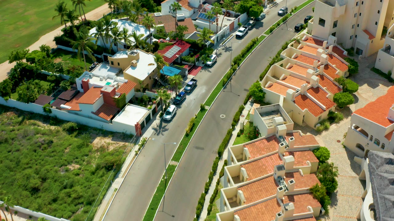 High aerial view in San Jose Del Cabo showing a curved residential road, cream homes with red-tiled roofs, parked cars, palms, green lots, and no visible people.