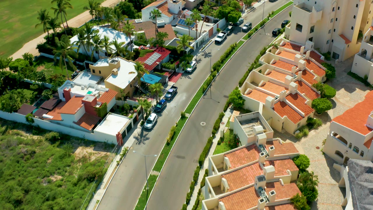 Overhead San Jose Del Cabo view showing a cream multi-story building, red-roof homes, a narrow road with cars, green landscaping, and no visible people.