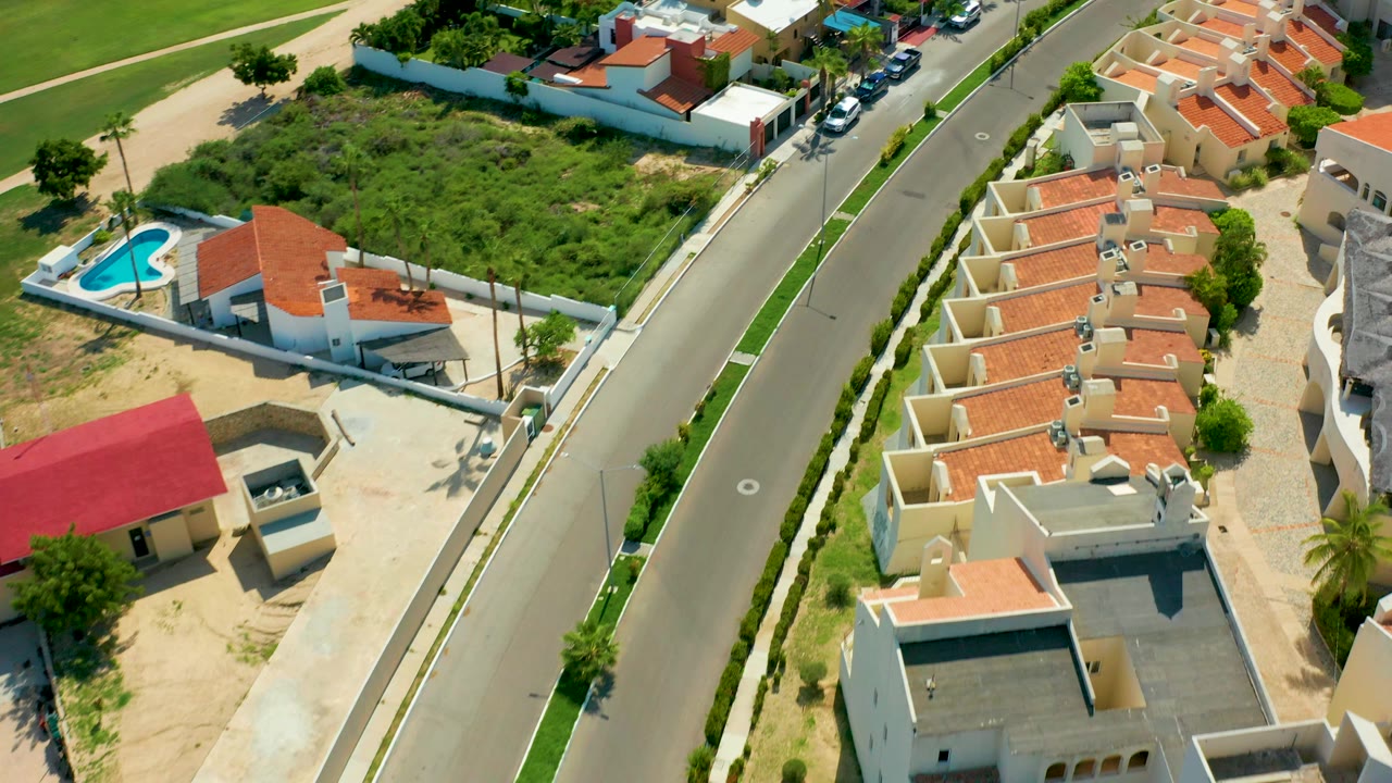 High aerial view over a San Jose Del Cabo residential street with red-tiled roofs, walled homes, a blue pool at left, green lots, parked cars, and no visible people.