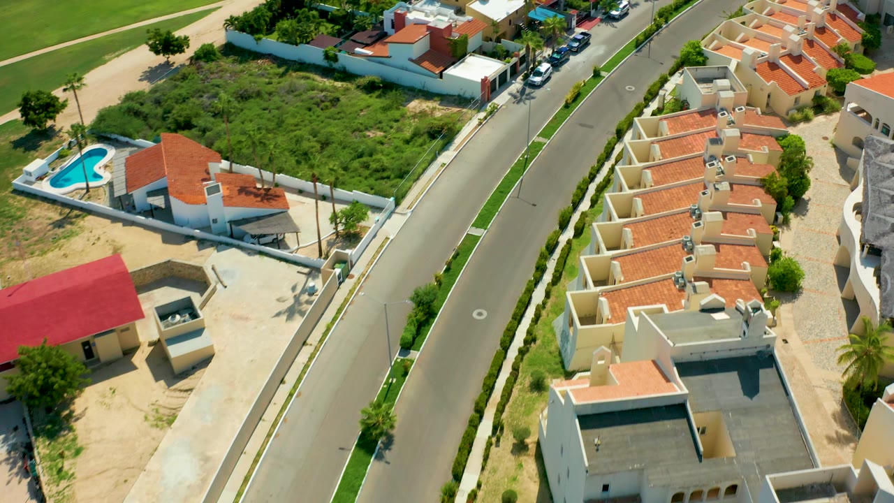 Aerial frame of San Jose Del Cabo homes lining a curved road, with red tile roofs on the right, a fenced green lot on the left, parked cars near the top, and no visible people.