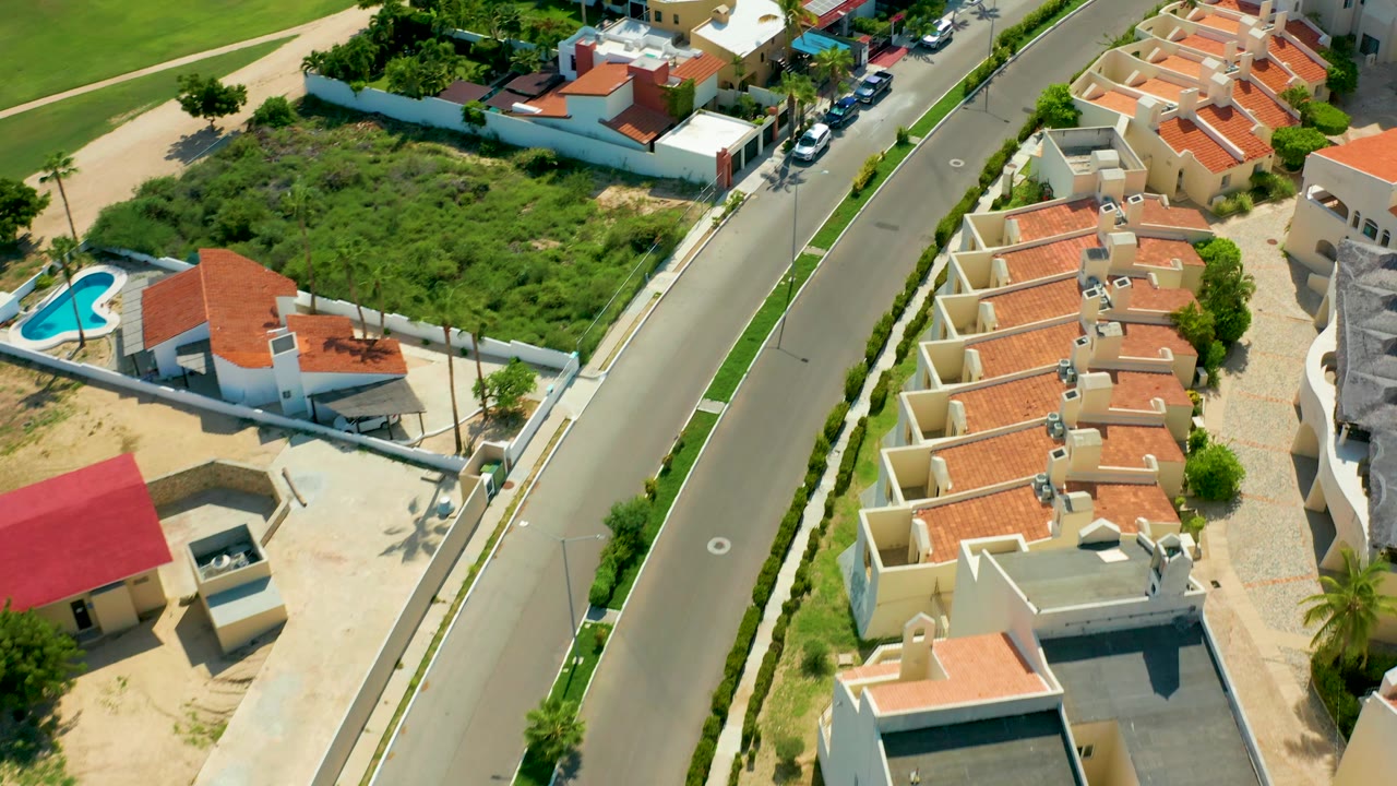 San Jose Del Cabo residential block seen from above with a blue pool in a walled property, red-roofed homes along the road, green vegetation, parked cars, and no visible people.