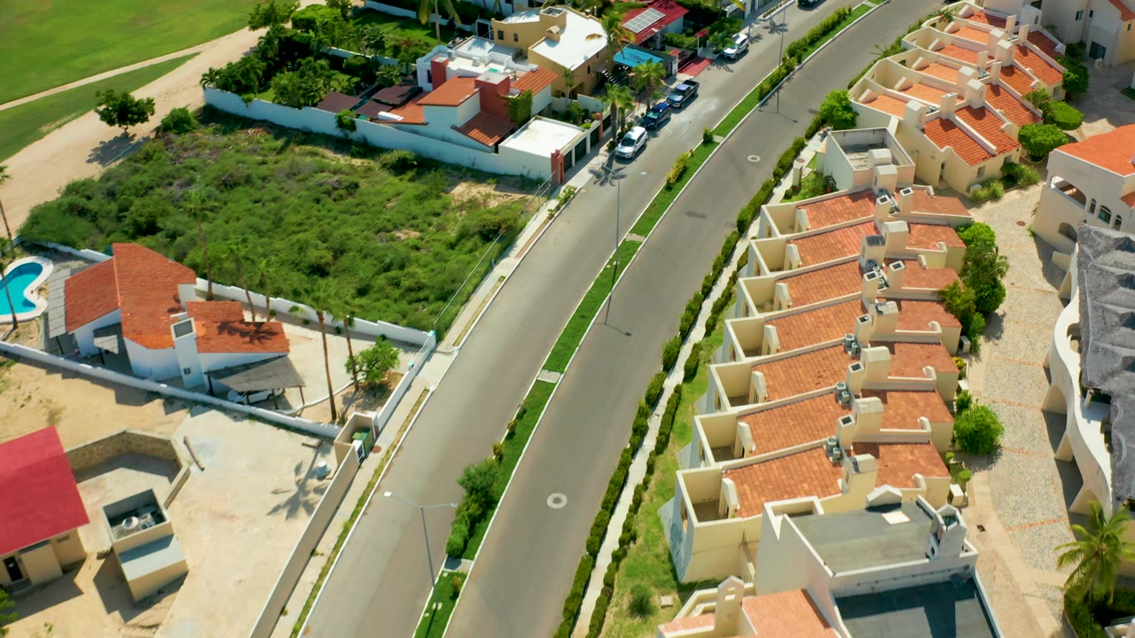 Residential street with a vacant green lot and rows of red tile roofs.