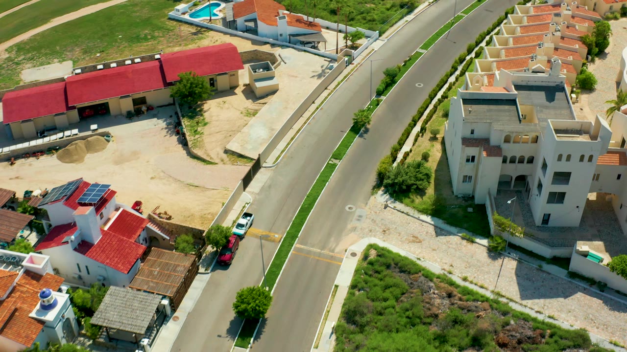 Drone view over a road beside a large cream house, red-roofed buildings, cars, and open lots.