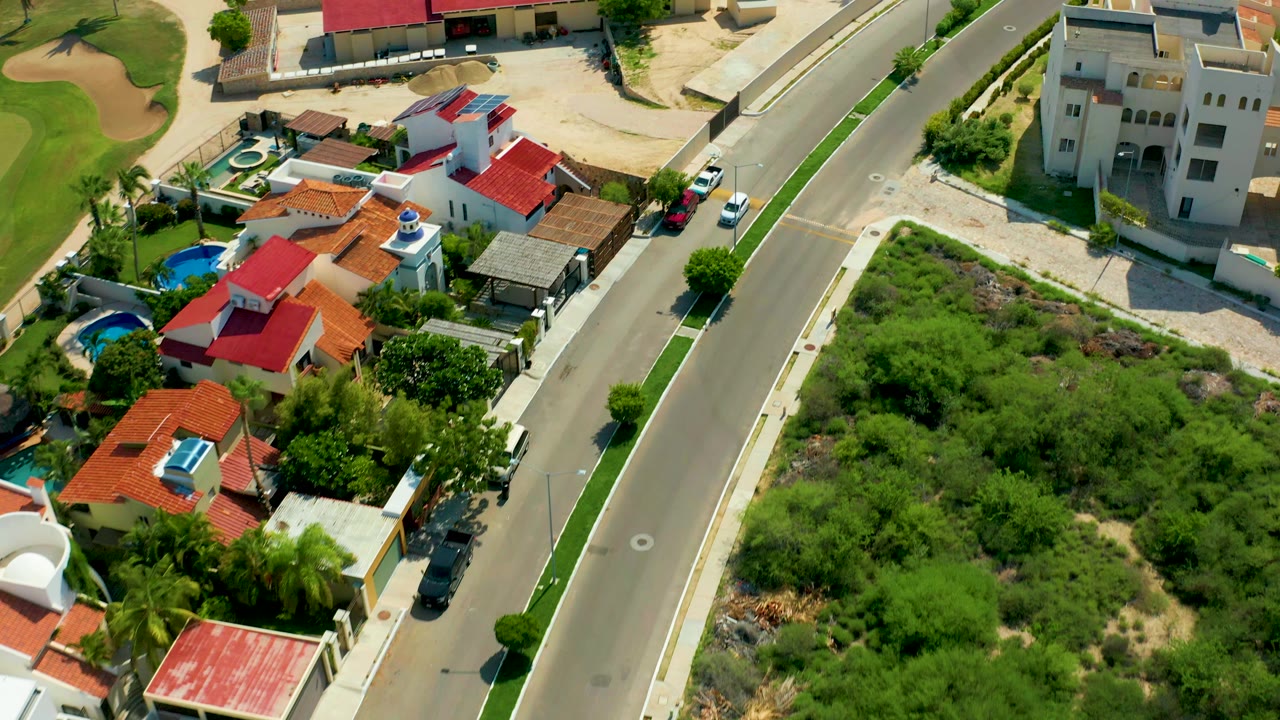 Aerial frame from San Jose Del Cabo showing a residential road beside red-roofed homes, trees, a blue pool, a green lot, and no visible people.