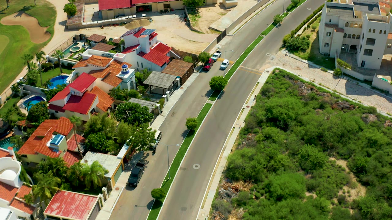 Aerial frame in San Jose Del Cabo with the road shifted toward center, red and cream houses, parked cars, green vegetation, a small pool, and no visible people.