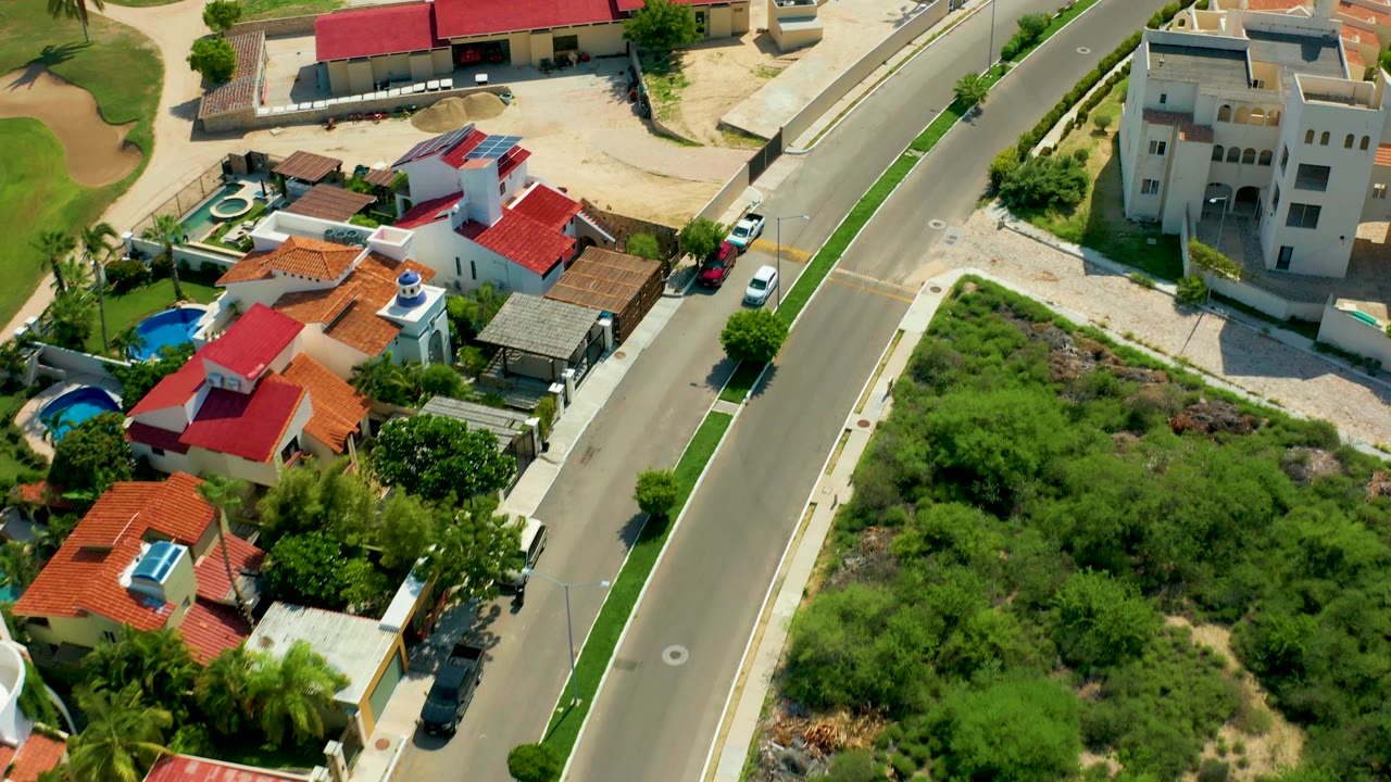 A San Jose Del Cabo aerial frame shows the residential street, red-roofed homes, a cream multi-story building, green open land, small pools, cars, and no visible people.