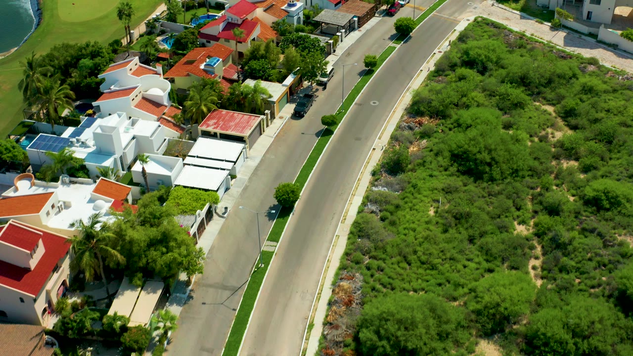 Aerial frame over San Jose del Cabo showing white homes with red-tiled roofs on one side of a paved residential road and dense green vegetation on the other, with no visible people.