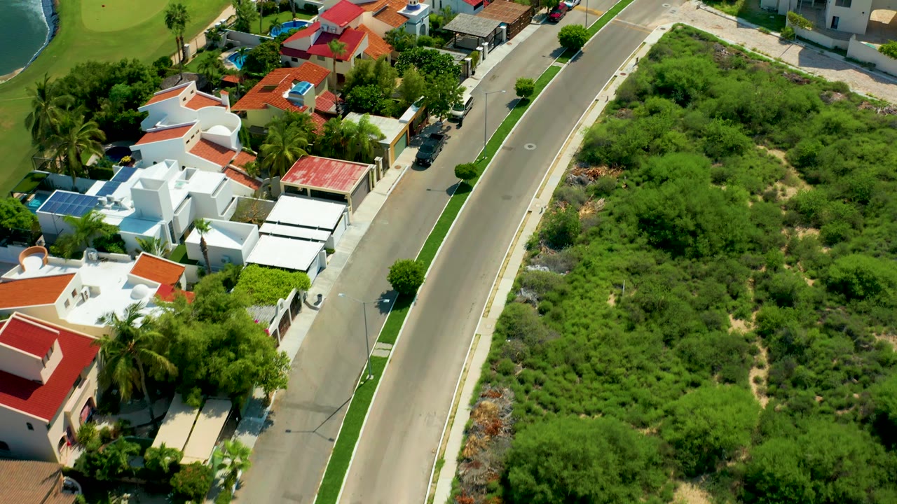 San Jose del Cabo aerial view slightly farther along the road, with white villas, red roof sections, parked cars near the curb, dense shrubs, and no visible people.