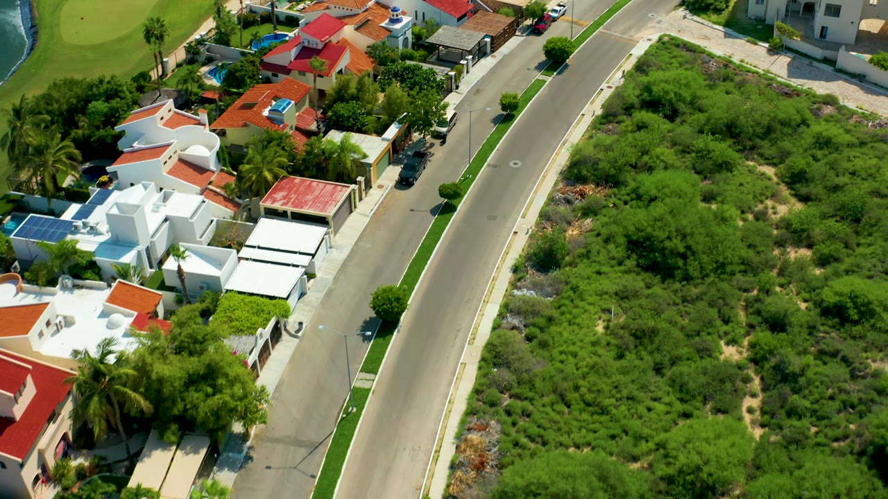 High drone frame in San Jose del Cabo with the paved road centered between white residential buildings, red-tiled roofs, a large green lot, and no visible people.