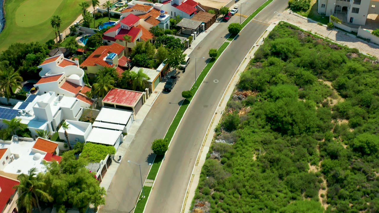 Overhead residential road view with white houses, red roofs, palms, and dense vegetation.