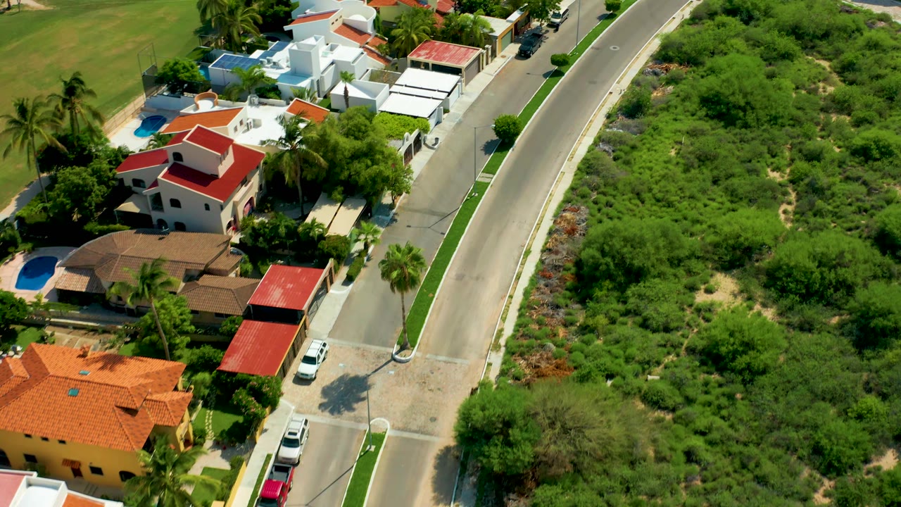 Aerial San Jose del Cabo frame showing red and white houses, a narrow residential road, cars near driveways, palm trees casting shadows, and no visible people.